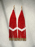 A pair of long beaded fringe earrings with red and white geometric beadwork and gold-tone hooks, displayed against a textured gray background.