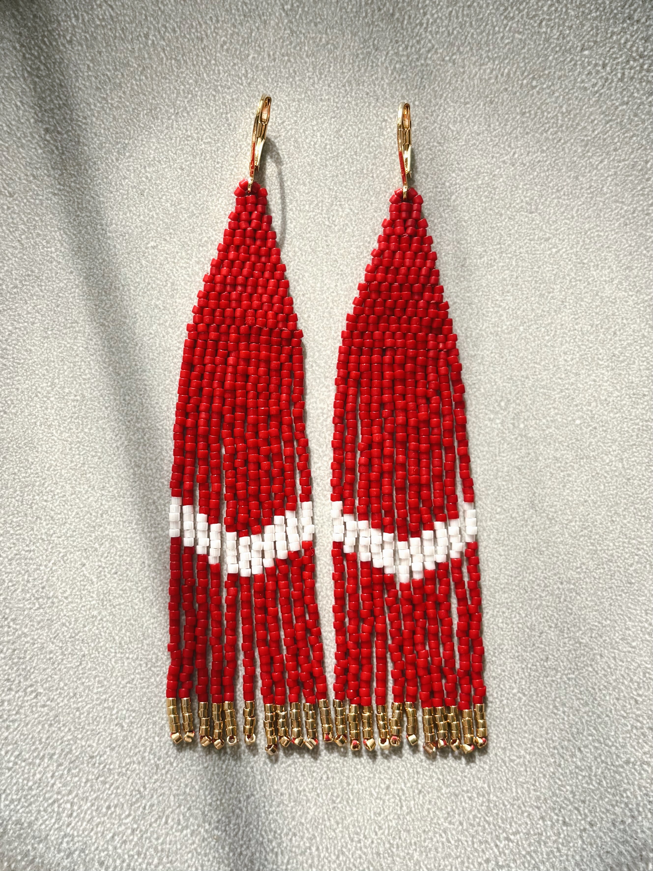 A pair of long beaded fringe earrings with red and white geometric beadwork and gold-tone hooks, displayed against a textured gray background.