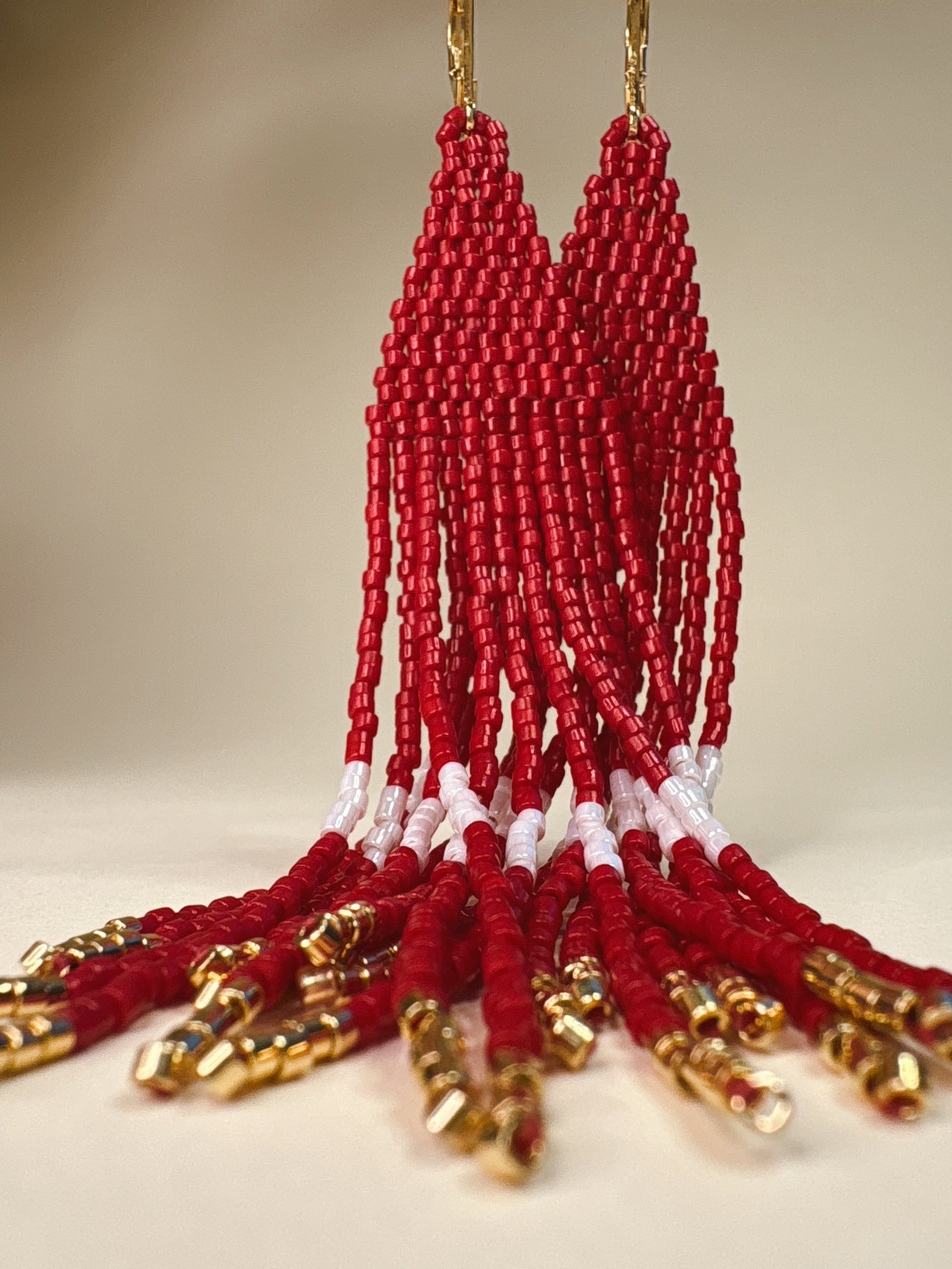 A pair of long beaded fringe earrings with red and white geometric beadwork and gold-tone hooks, displayed against a textured gray background.