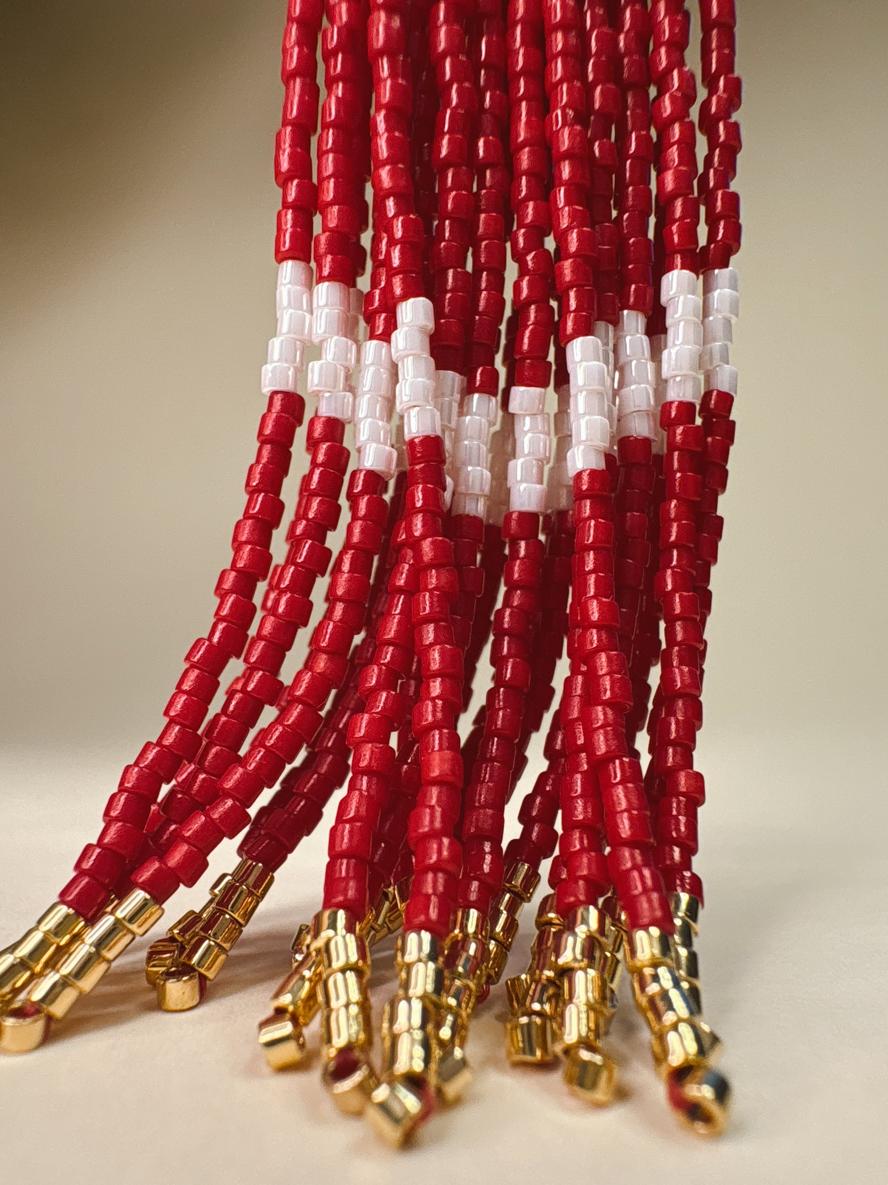 Close-up of multiple strands of red and white beaded tassels with gold metal tips, arranged against a plain light background.
