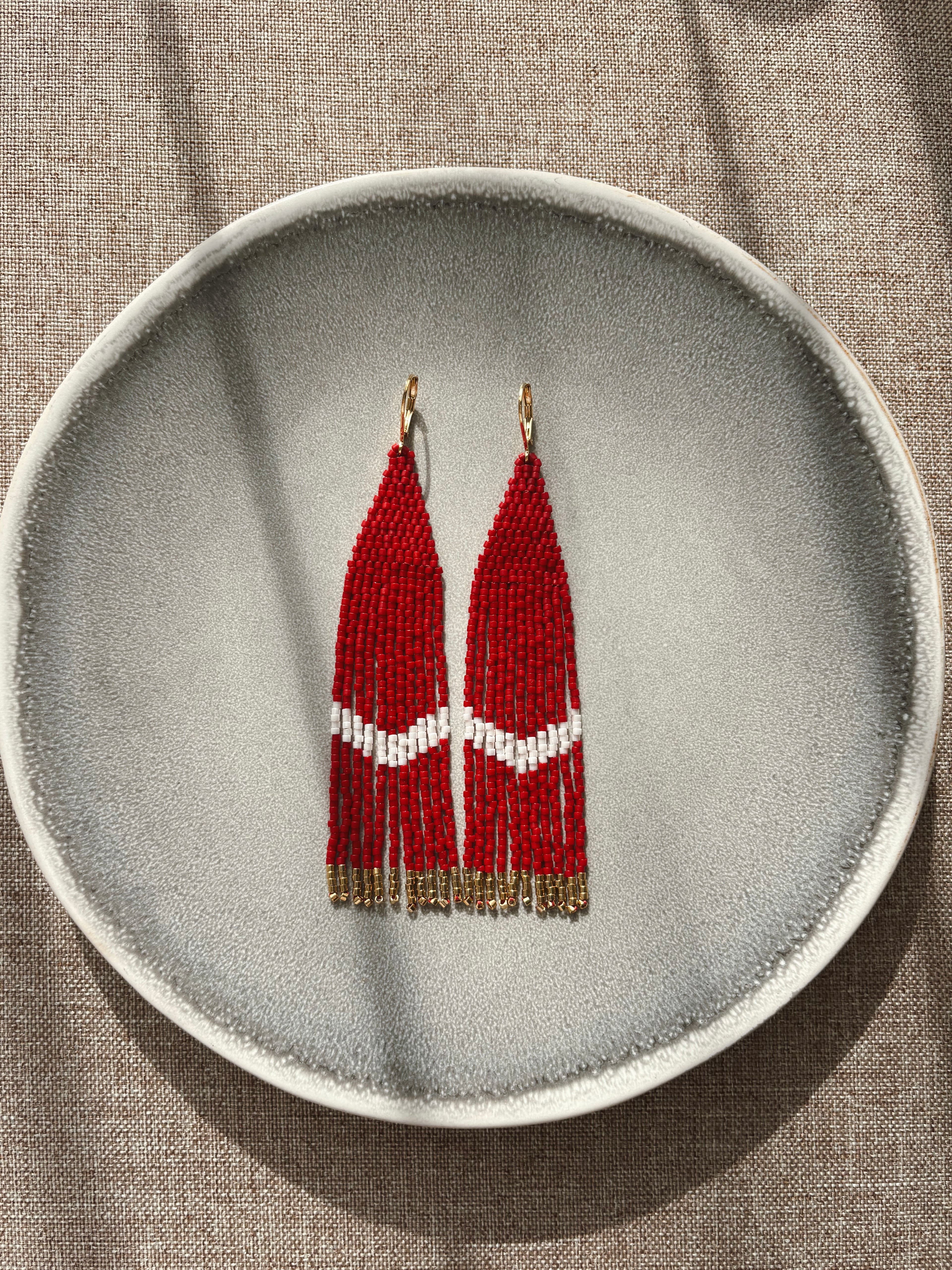 A pair of long beaded fringe earrings with red and white geometric beadwork and gold-tone hooks, displayed against a textured gray background.